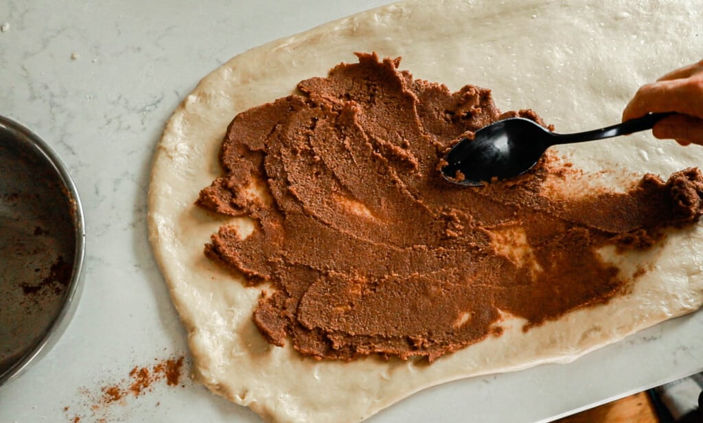 cinnamon sugar butter mixture being spread out onto rolled out dough with a spoon on a white countertop