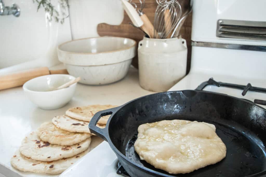sourdough flatbread frying in a cast iron skillet with olive oil. More flatbreads are layered on the white countertop