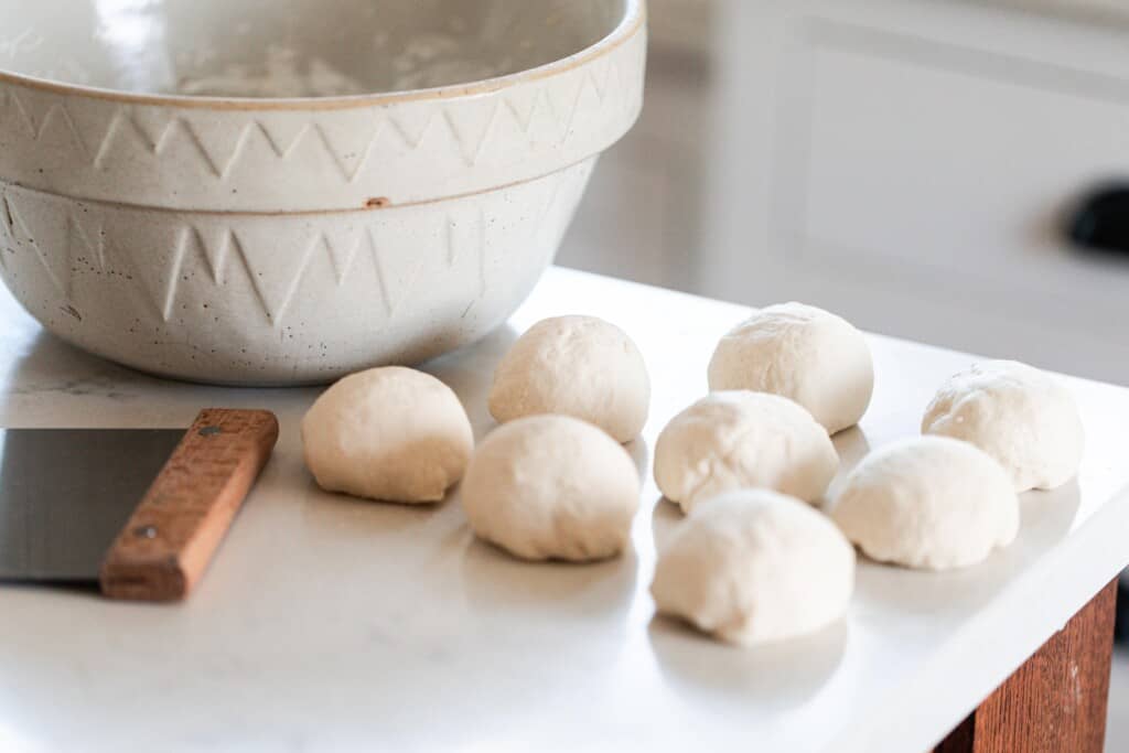 sourdough flatbread dough rolled into balls on to a white quartz countertop with a bench scraper to the left and a large cream colored bowl in the back