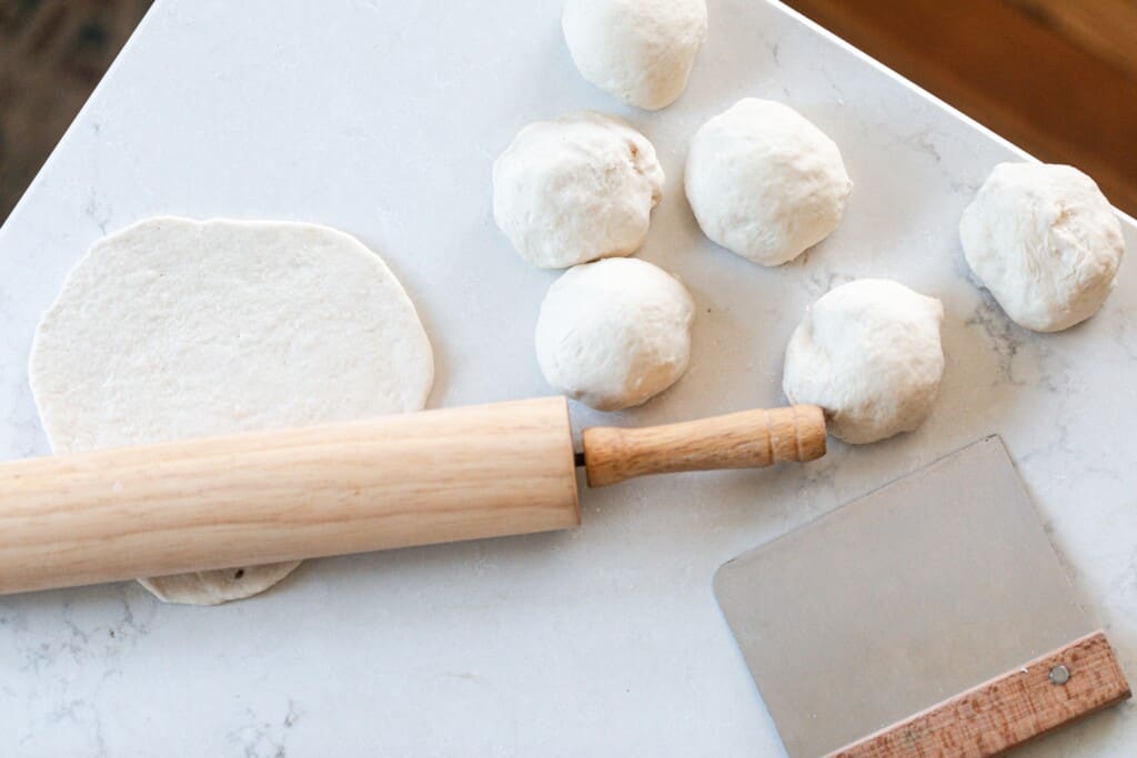 sourdough flatbread dough being rolled out with a rolling pin on a white quartz countertop with dough balls to the right and a bench scraper