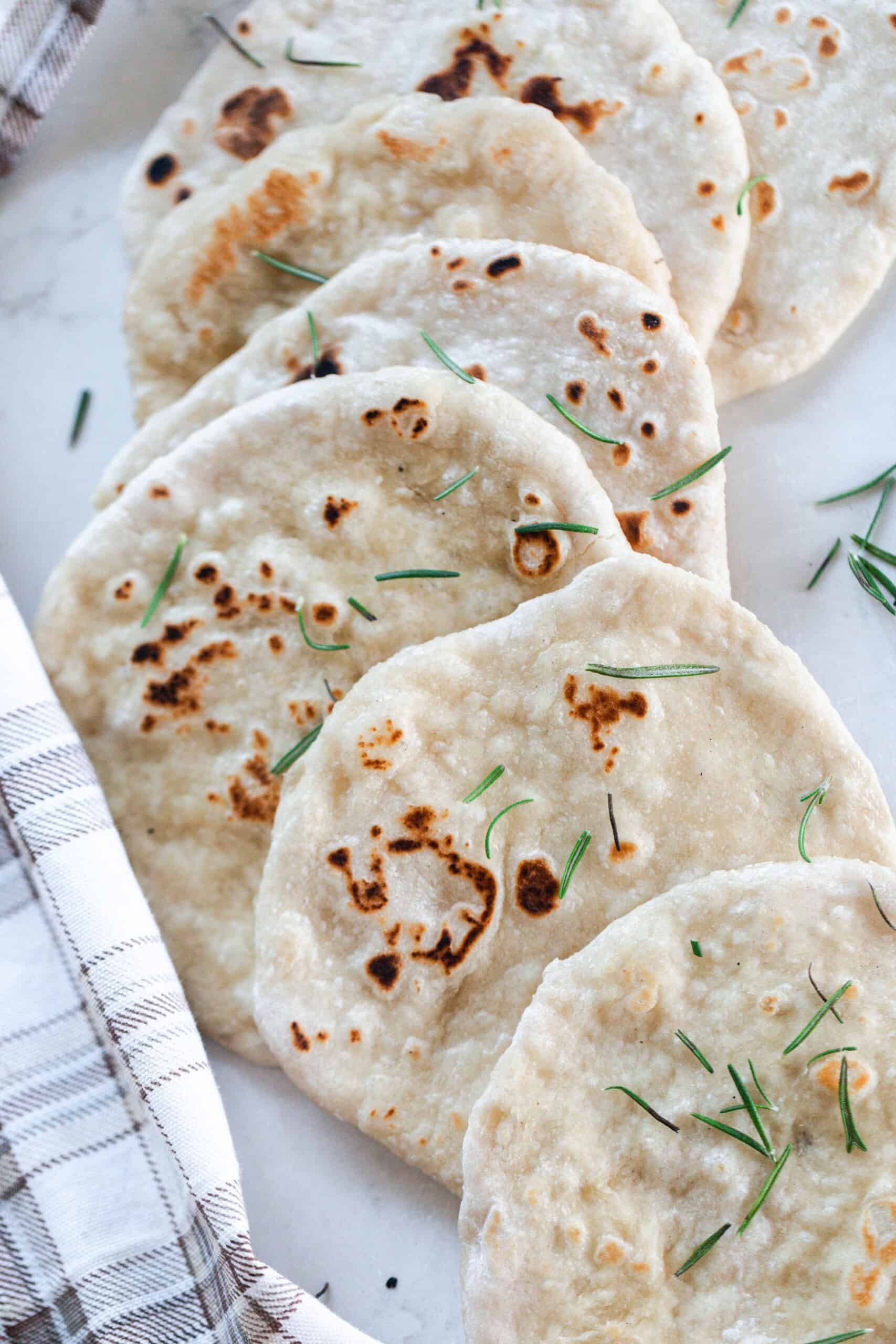 sourdough flat bread topped with fresh rosemary layered in a line on a white quartz countertop with a plaid towel to the left