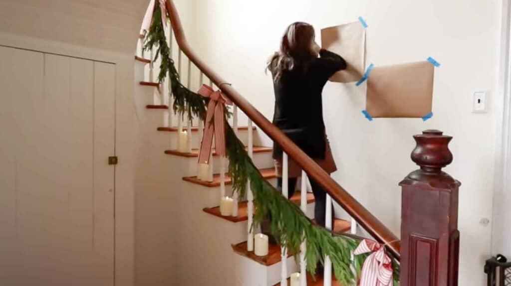woman on a staircase taping up brown paper with painters tape onto a wall to map out where she wants pictures to be hung
