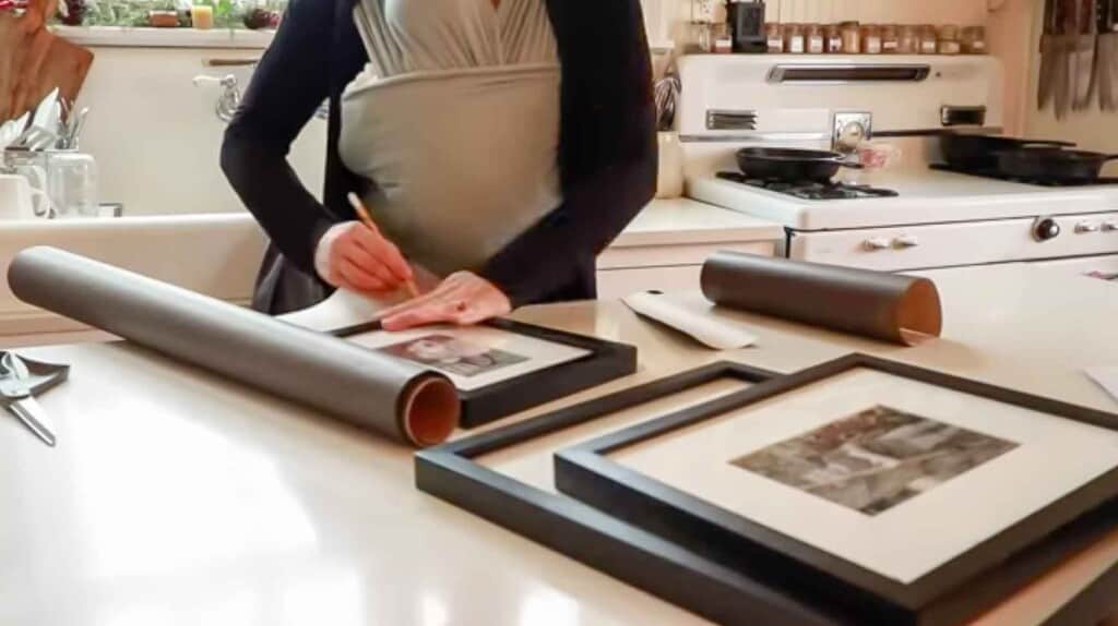 woman wearing a baby in a wrap tracing out picture frames on brown paper in her white kitchen