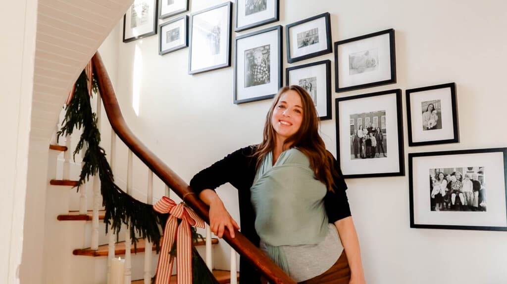 woman wearing a baby standing on her farmhouse staircase with a staircase gallery wall with family pictures hung in black frames. the staircase is wrapped wit greenery and red and white stripped bows