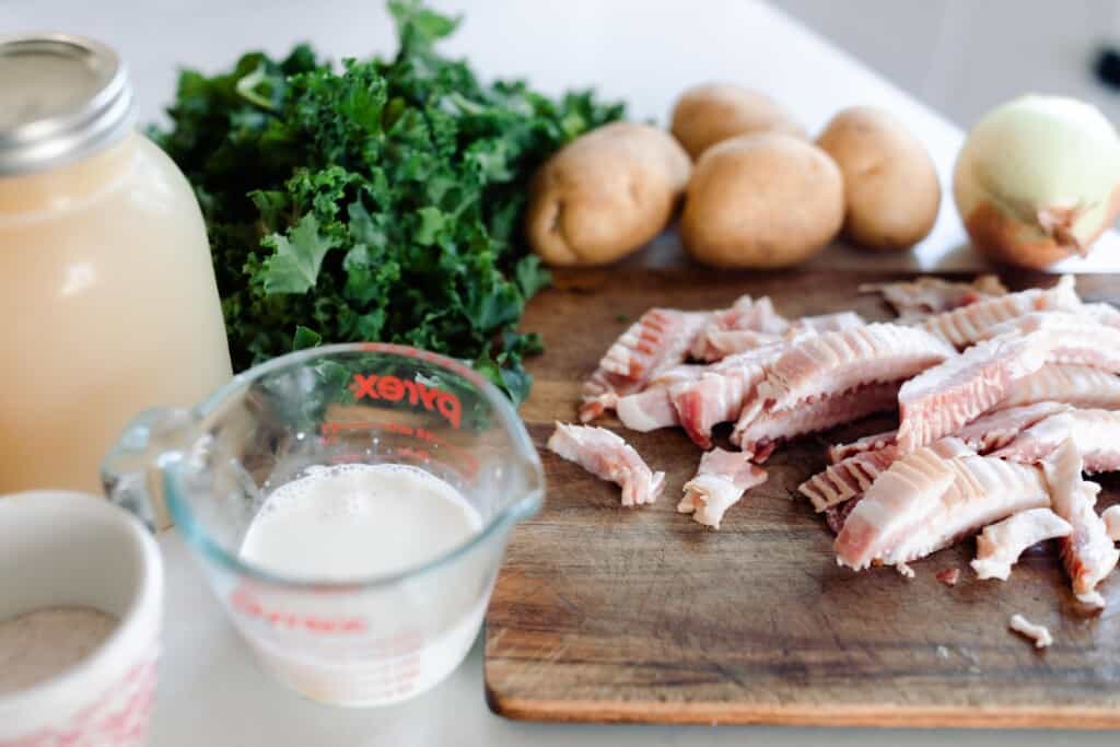 wooden cutting board with slices of bacon on it. Potatoes, kale, chicken broth in a mason jar, and a measuring cup of cream on a white quartz counter