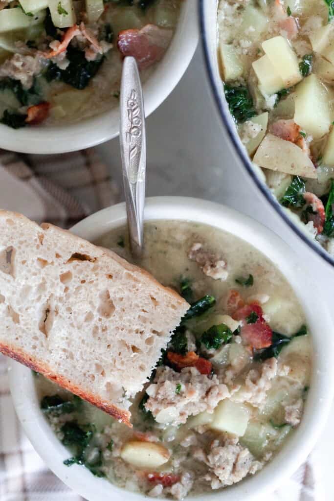 overhead photo of two bowls of zuppa toscana soup with a slice of homemade sourdough bread sitting on the rim of the white bowl and a pot of soup to the right
