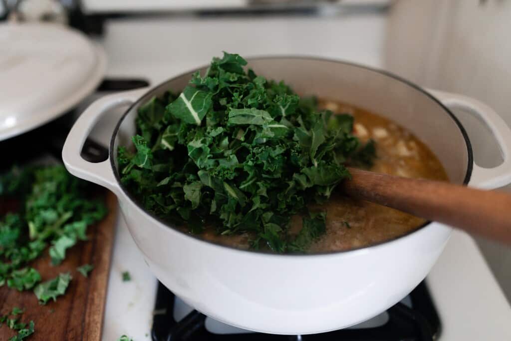 de-stemed and chopped kale being added to a white dutch oven full of soup on a white stove