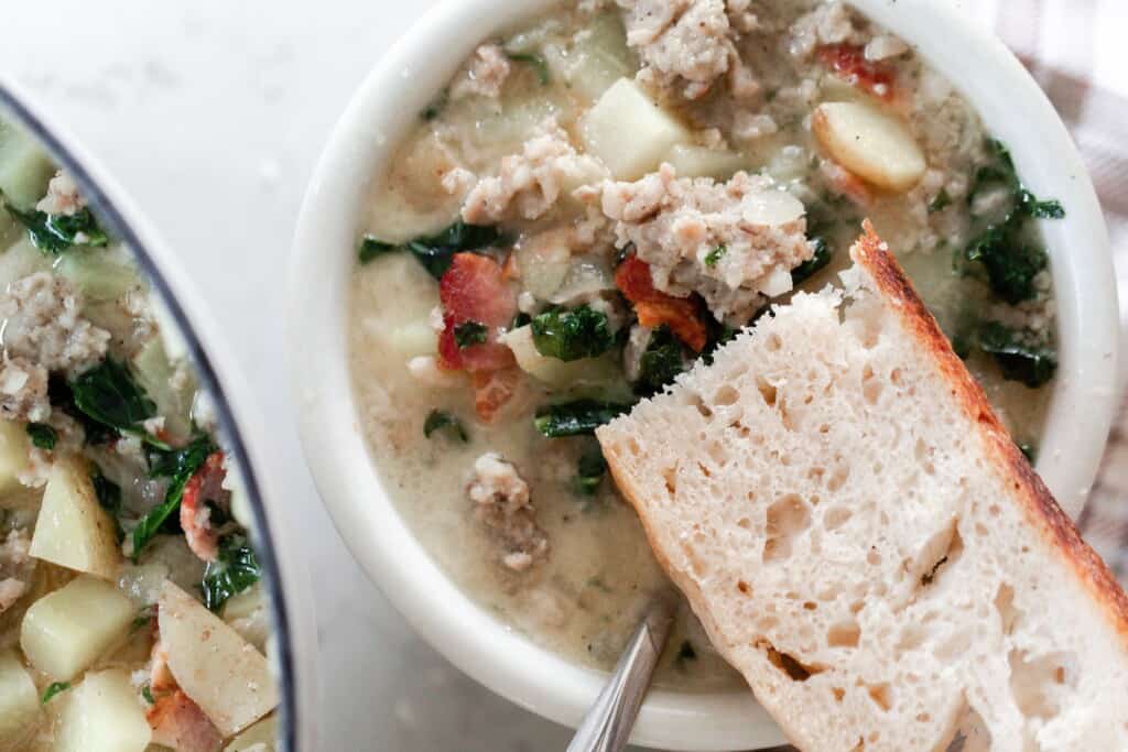 overhead photo of a white bowl of creamy zuppa toscana soup with a slice a sourdough bread resting on the bowl of the rim with the corner of the soup pot to the left
