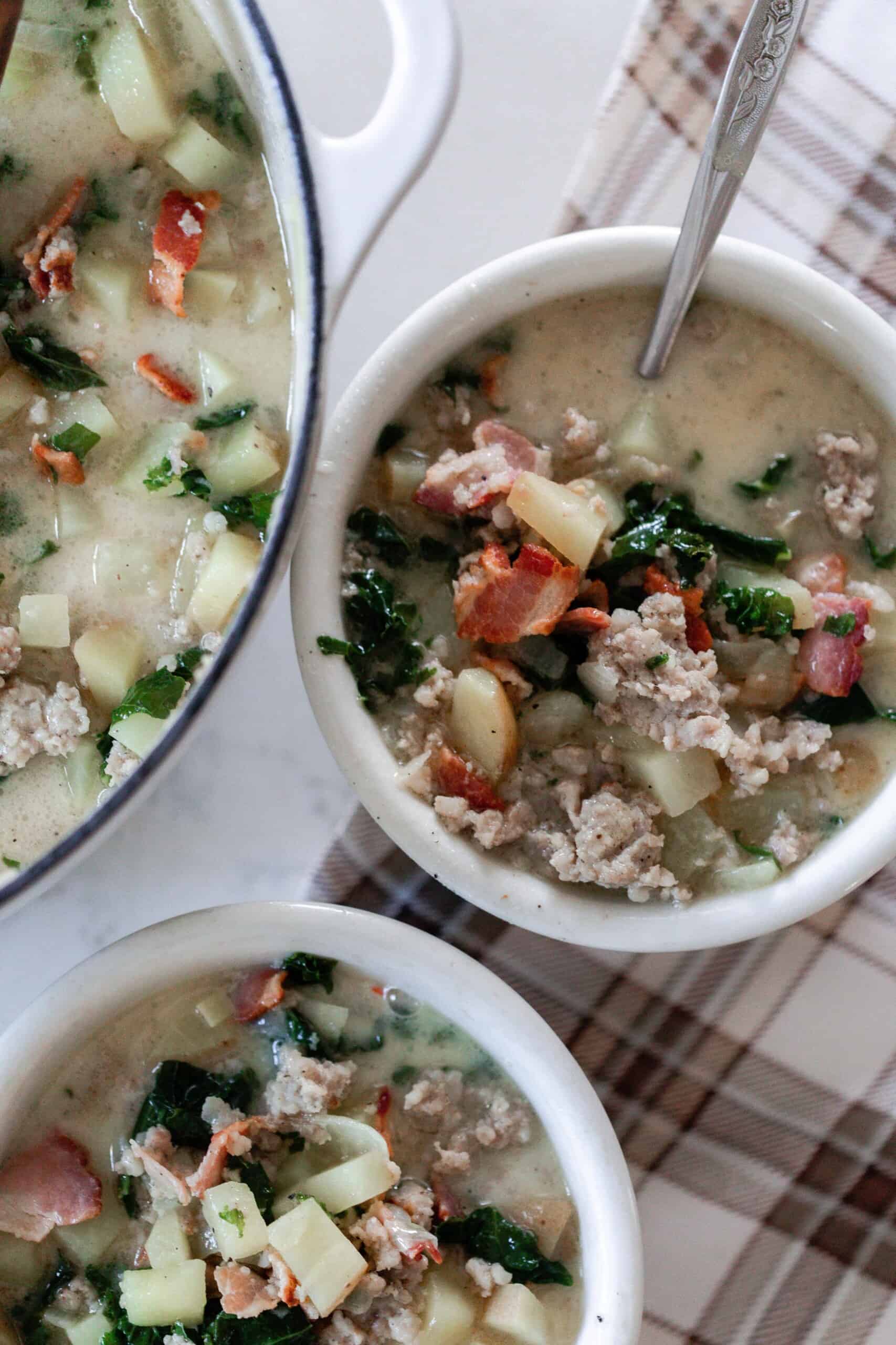 two bowls of cuppa toscana soup with potatoes, sausage, bacon, kale in white bowls with a pot of soup to the left.