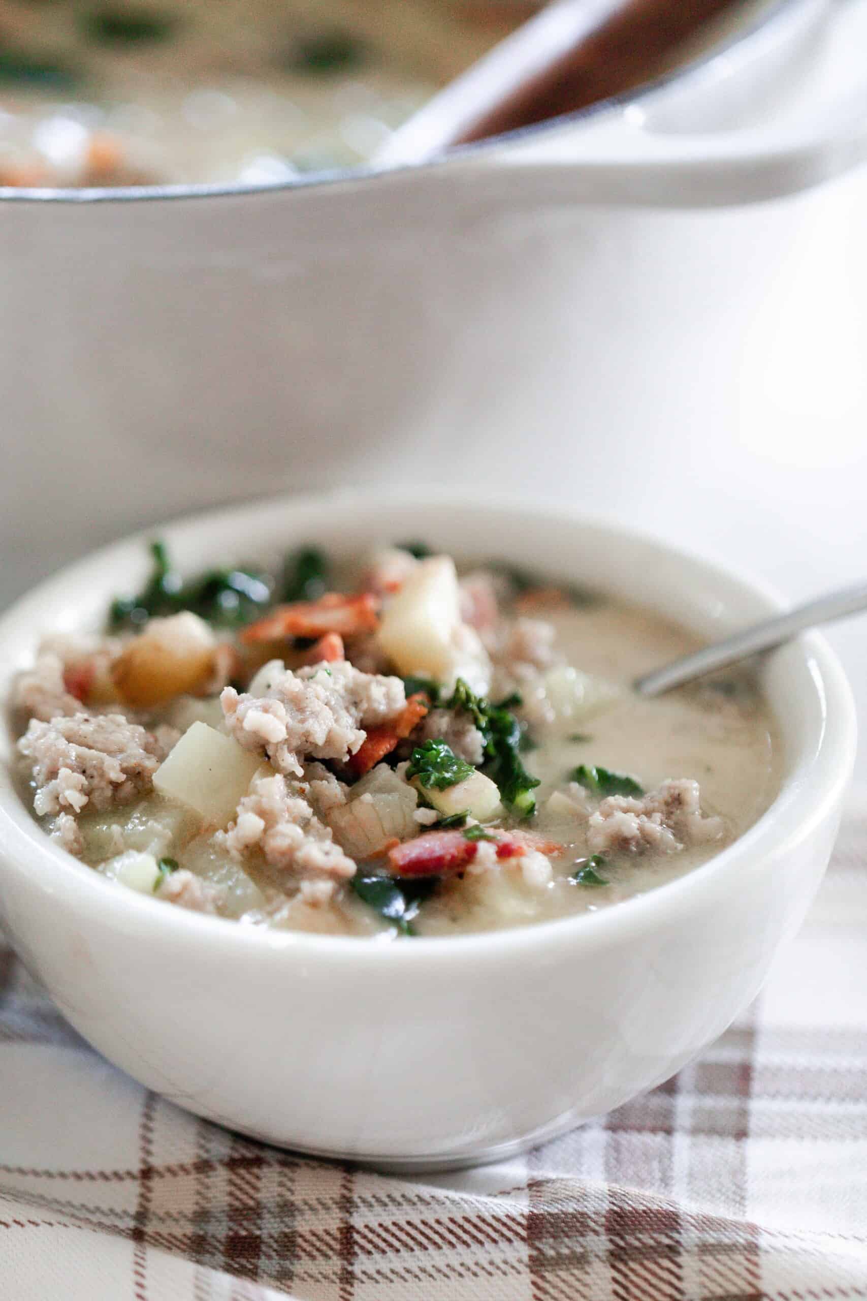 bowl of zuppa toscana soup with a spoon on a cream and brown plain napkin with a white dutch oven in the background