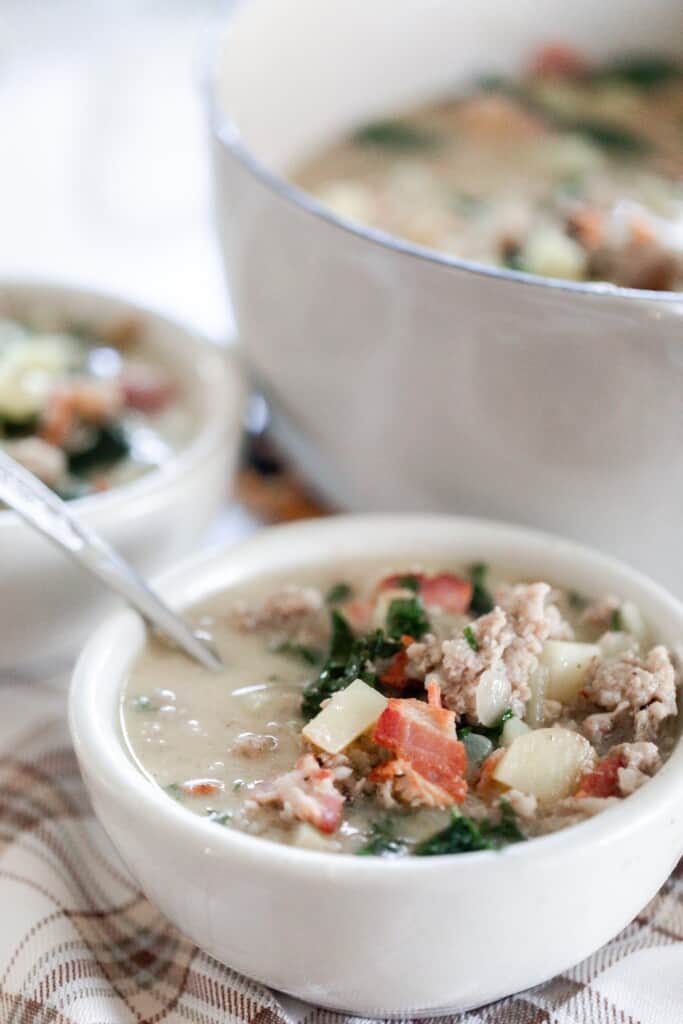 white bowl of easy zuppa Toscana soup with potatoes, sausage, bacon, and kale with a large white dutch oven in the background