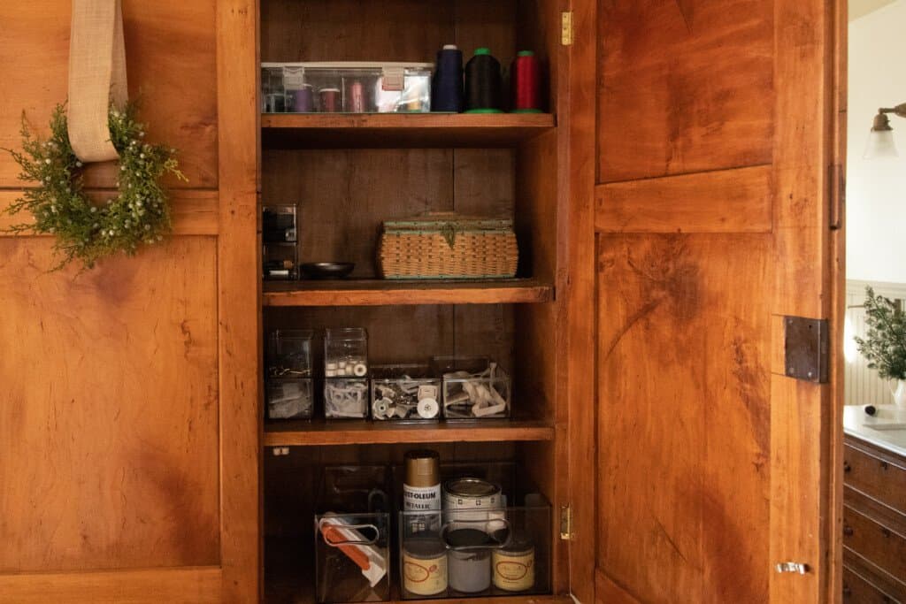 A wooden antique cabinet with the right door opened revealing organized sewing material. Spools of thread are stacked on the top shelve, clear bins of bobbins, sewing feet, etc in the middle, and pain and other supplies in clear bins on the bottom shelve