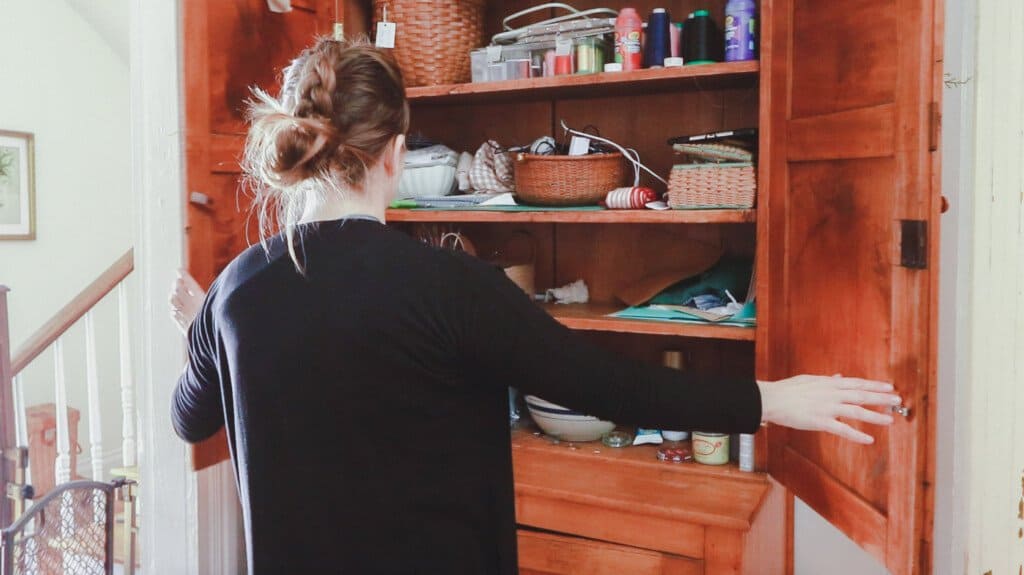 a woman opening up a craft cabinet doors to reveal an unorganized space with thread, baskets, fabric, and other craft items disorganized on shelves.