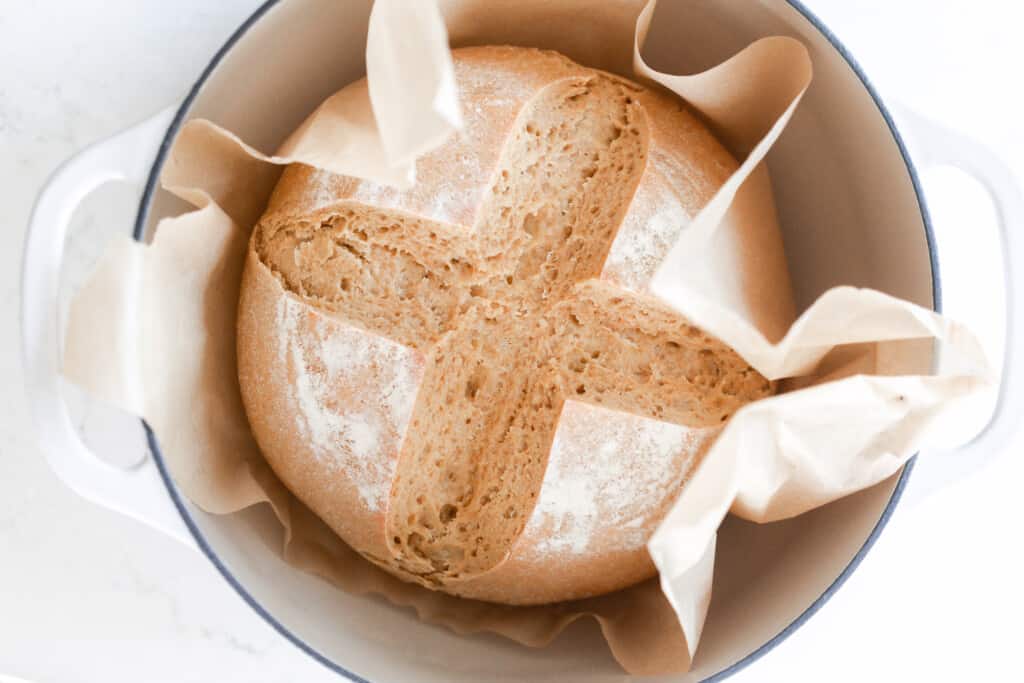 overhead photo of einkorn sourdough bread with a cross pattern scorned on top in a parchment paper lined ditched oven