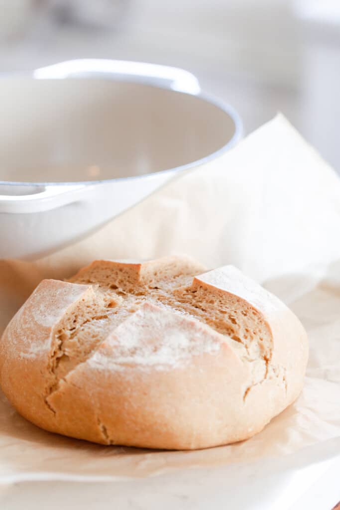 A loaf of sourdough einkorn bread with a cross shape scored on the top resting on parchment paper with a white dutch oven in the back