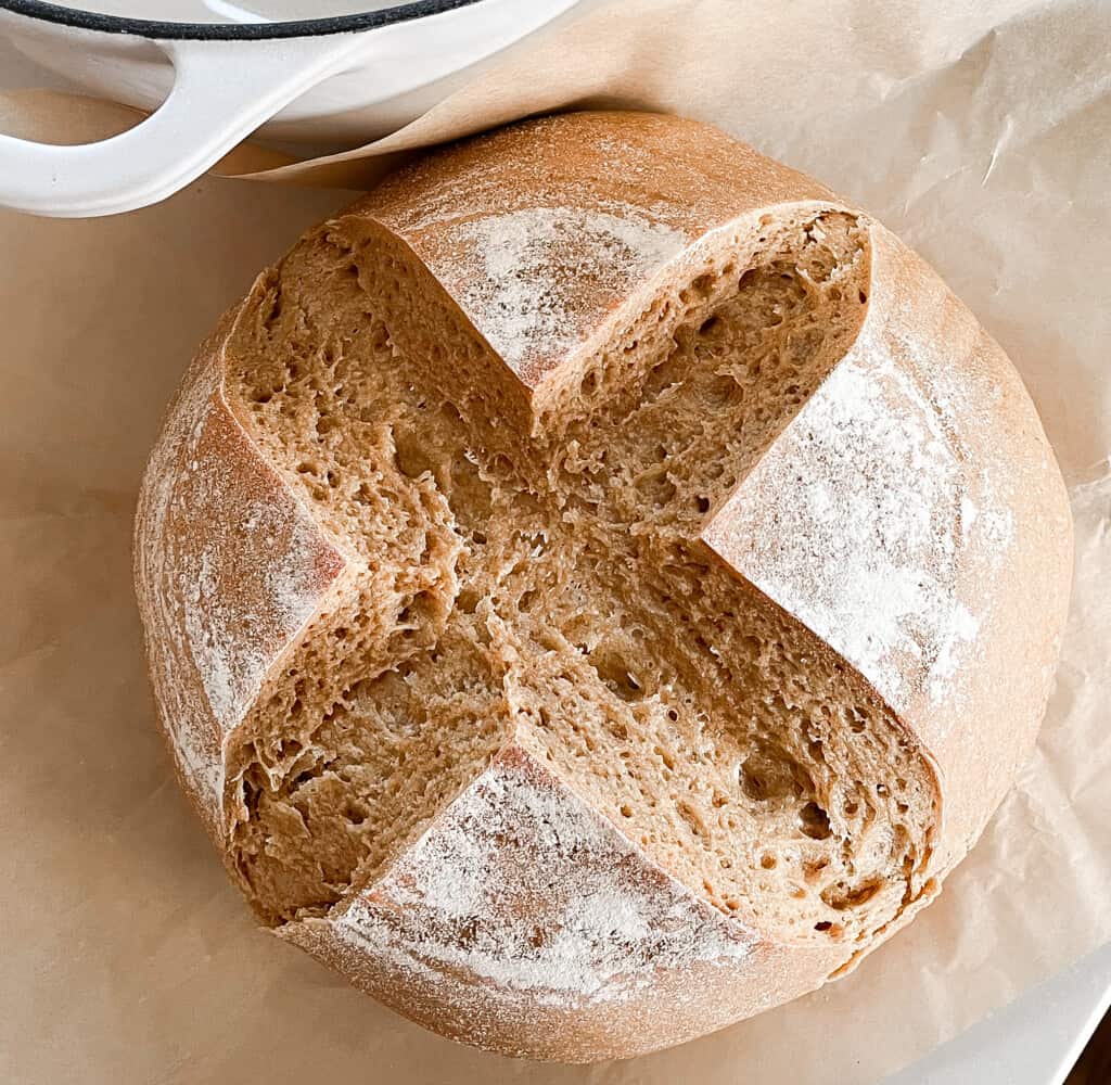 overhead photo of a round loaf of einkorn sourdough bread with a cross pattern scored. The loaf sits on a parchment with a white dutch oven right behind it.