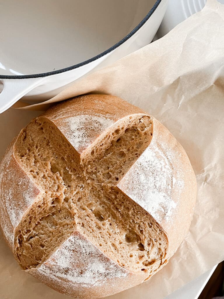 overhead photo of a loaf of sourdough einkorn bread on parchment paper on a white quartz countertop with a white dutch oven in the back left corner.