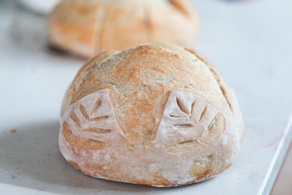 side view of a loaf of sourdough bread scored with a cross and wheat patterns on resting on a white countertop