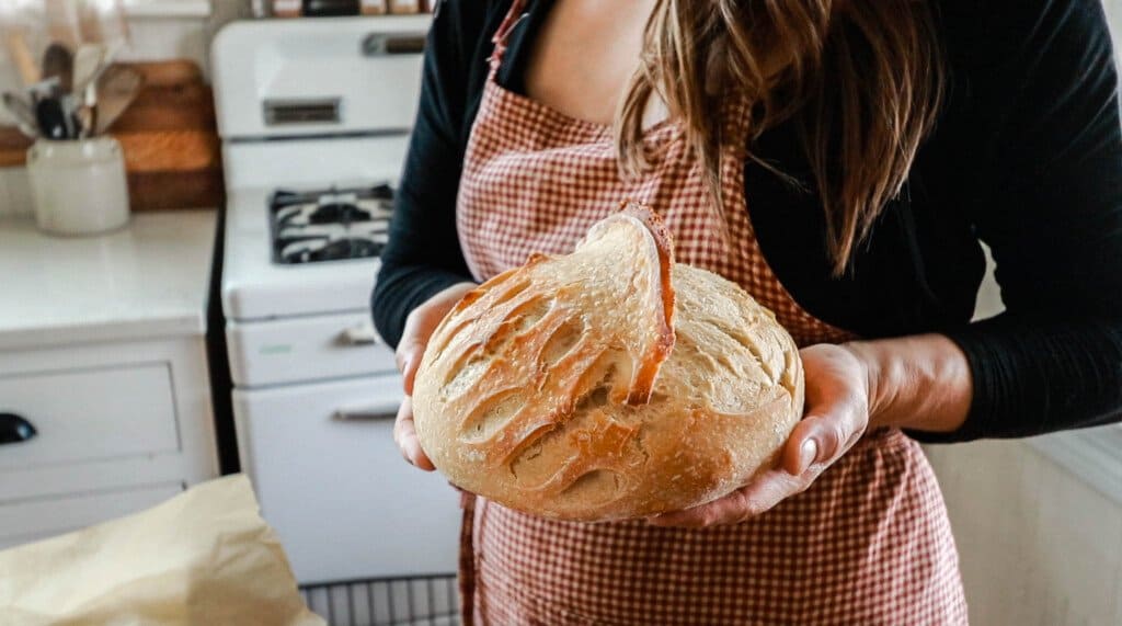 a woman wearing red apron holding a loaf of sourdough bread in her white kitchen