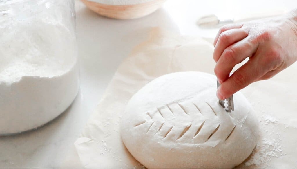 handing using a razor to score sourdough bread dough on parchment paper on a white countertop