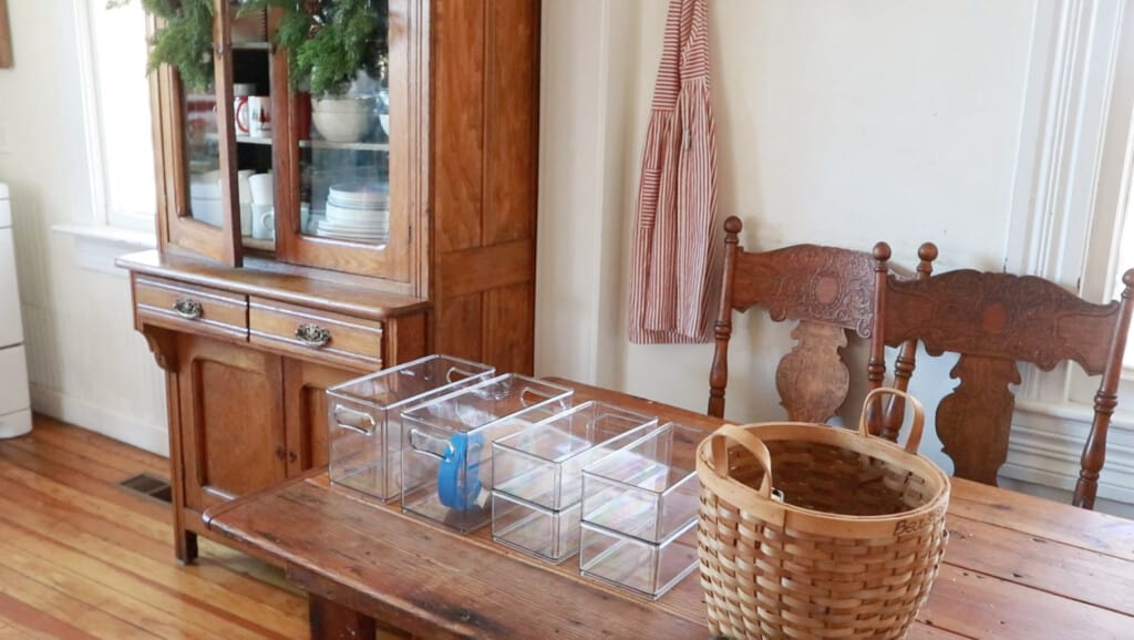 clear plastic organizational bins on a wooden kitchen table with a basket to the right. An antique chair and a red and white checked apron hanging on the wall are in the background