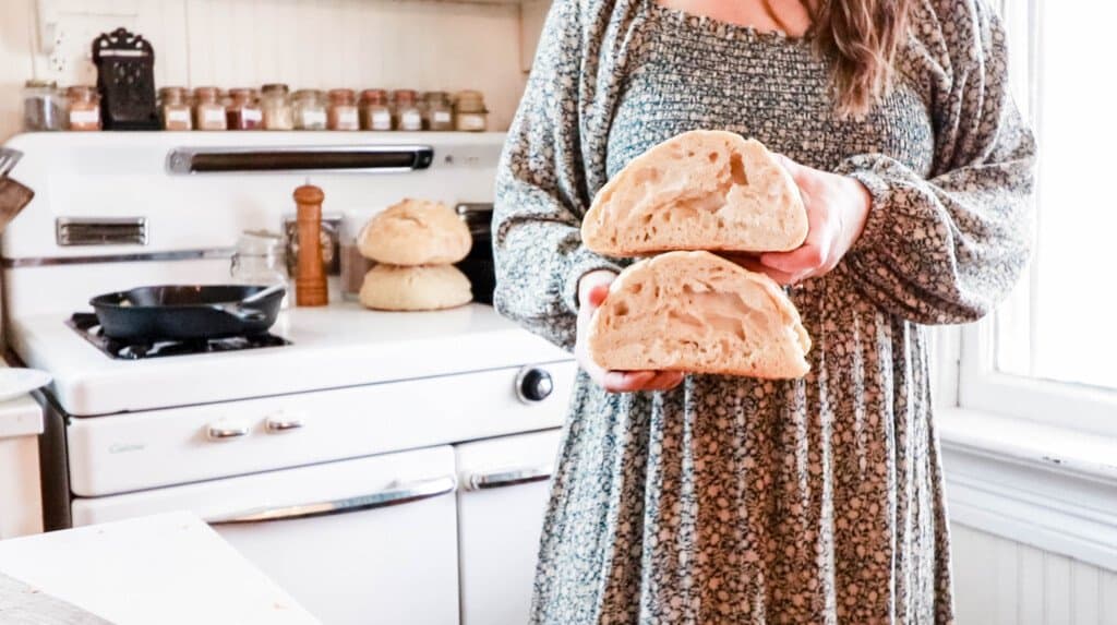 woman wearing a green floral dress holding a loaf of sourdough bread cut in half. A white vintage stove is in the background