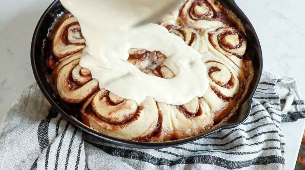 cream cheese icing being poured over sourdough cinnamon roll sin a cast iron skillet ton a white and black stripped towel
