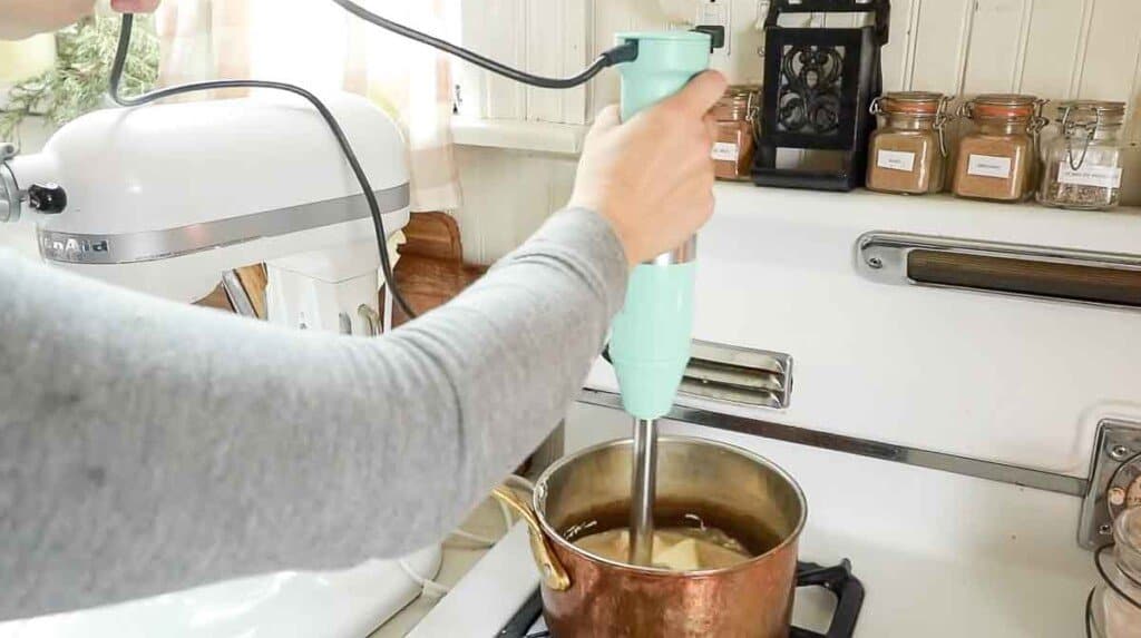 teal immersion blender combing cream cheese, maple syrup, cream, and vanilla in a copper pot on a vintage stove