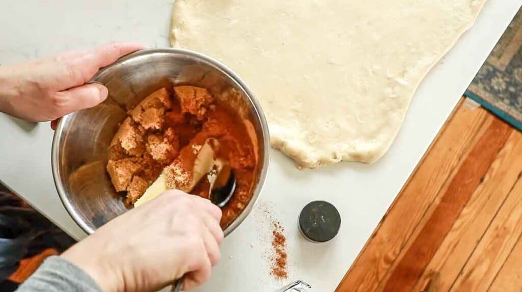 Hands holding a spoon mixing together butter, cinnamon, and brown sugar in a metal bowl. Dough rolled out into a rectangle