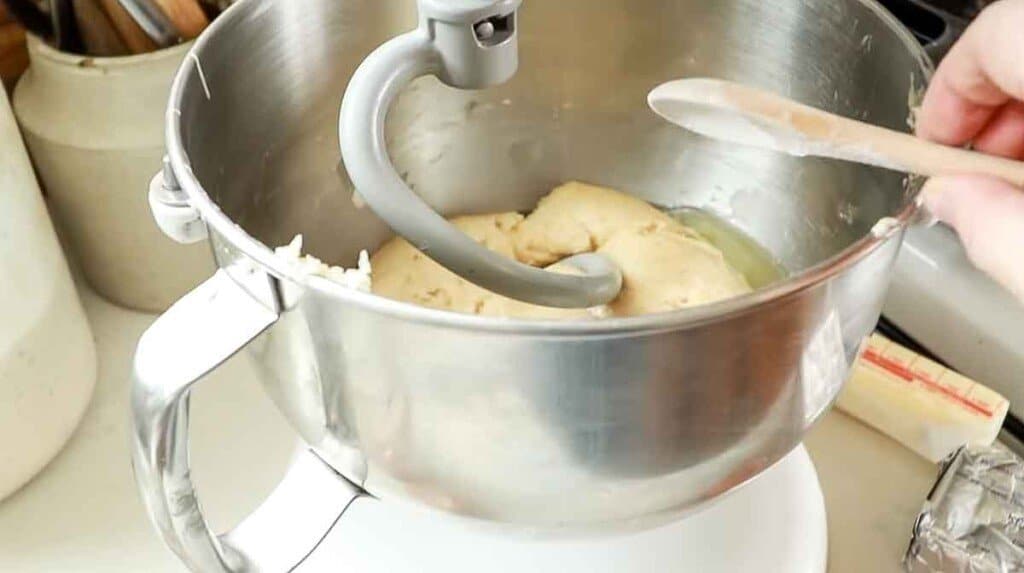 a wooden teaspoon with making soda being added to dough in a stand mixer with a dough hook