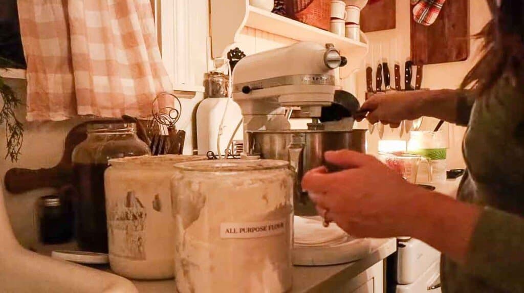 flour in a black measuring cup being added to a Kitchenaid stand mixer with jars of flour and sourdough starter to the left