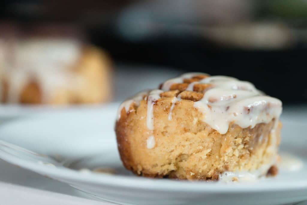 side view of a cinnamon roll made with einkorn flour and topped with cream cheese frosting on a white plate. Another white plate with a cinnamon roll in the background