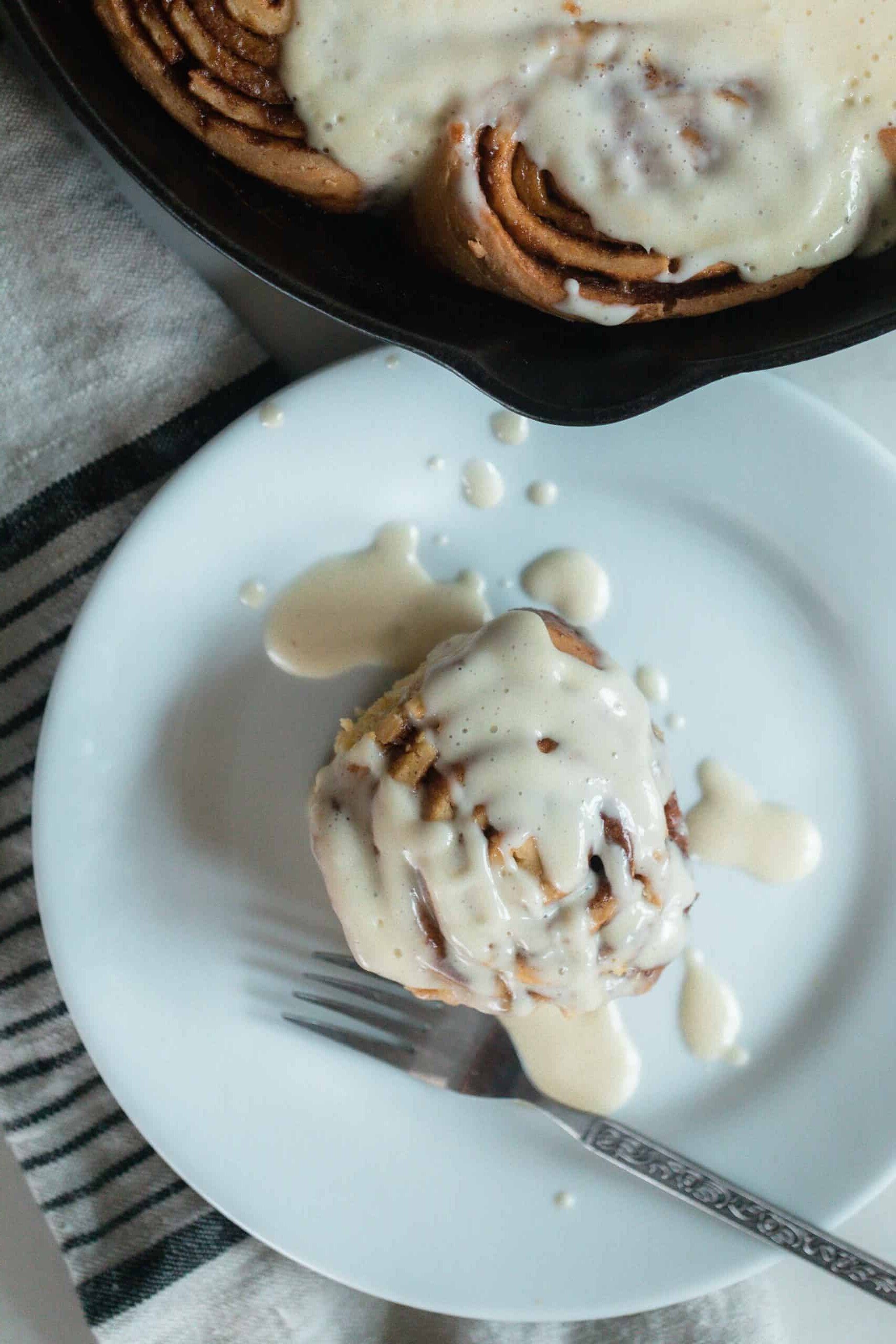 overhead photo of a einkorn cinnamon roll topped with cream cheese frosting on a white plate with a fork. A cast iron skillet with more rolls is behind the plate