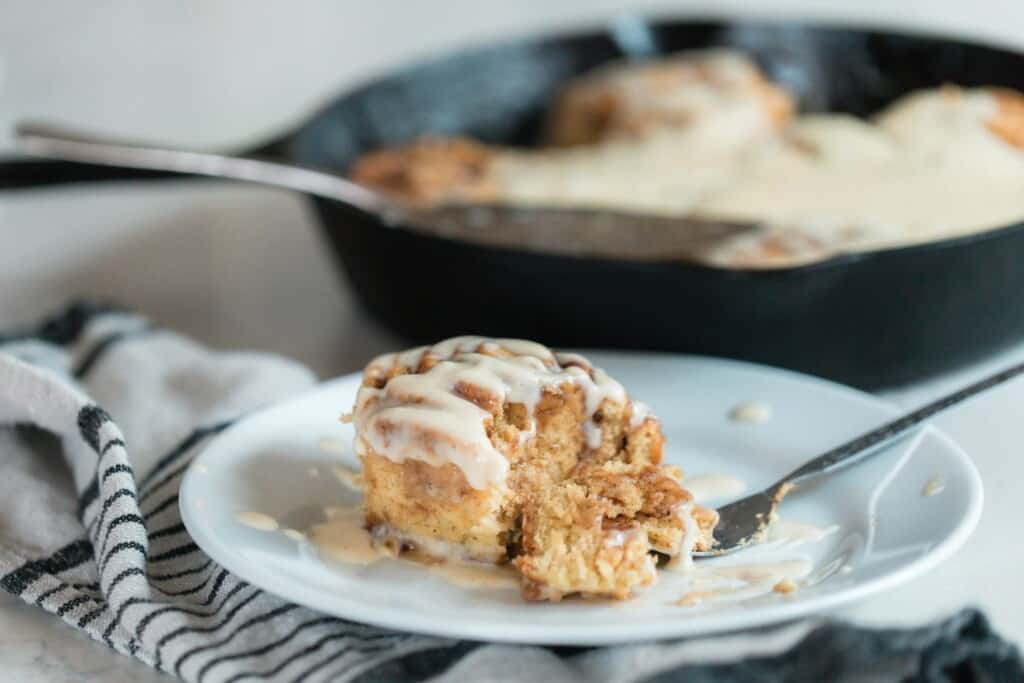 einkorn cinnamon roll on a white plate with a fork taking a forkful out. The plate sits on a black and white towel and a cast iron skillet full of cinnamon rolls is in the background