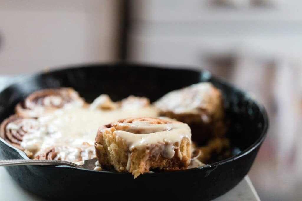 einkorn cinnamon roll being lifted out of a cast iron skillet on a white countertop with a vintage stove in the background