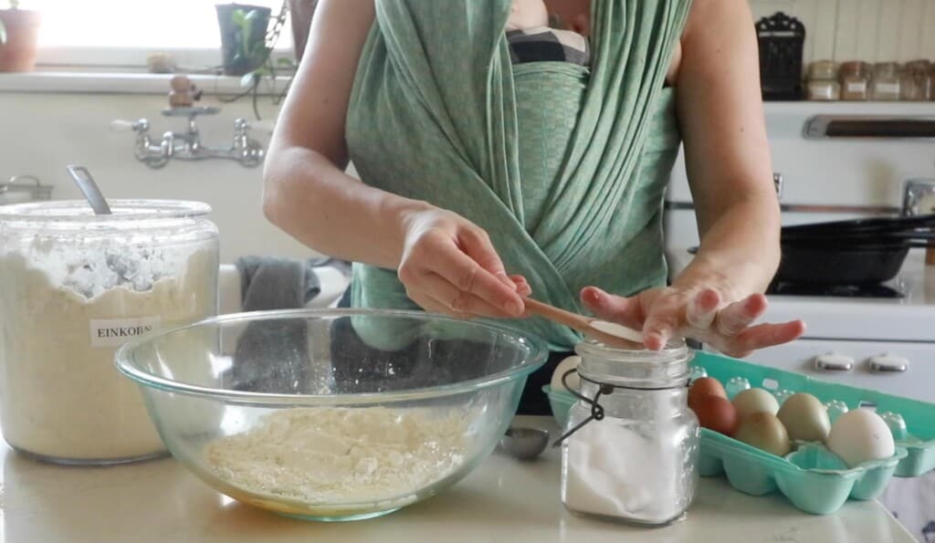 a women wearing a baby in a green wrap adding salt to flour and eggs in a glass bowl