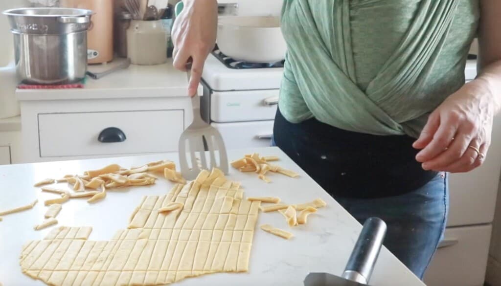women using a metal spatula to scrape off sliced noodles off of a white countertop