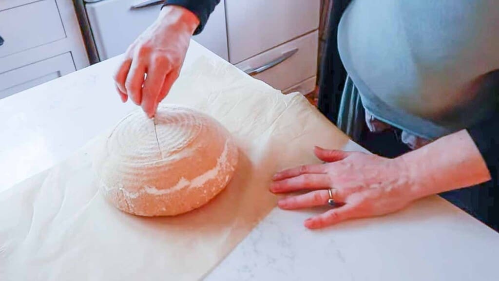 scoring a loaf of einkorn bread on a sheet of parchment paper on a white quartz countertop