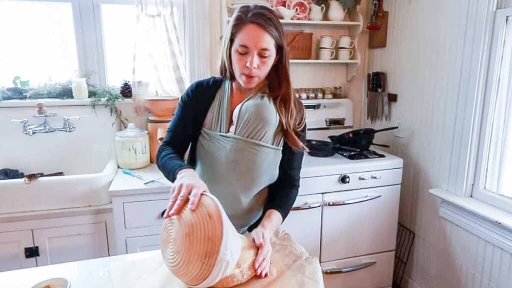 woman wearing a baby taking einkorn sourdough bread dough out of a banneton basket and placing it on a sheet of parchment paper