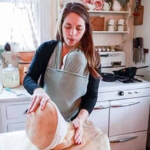 woman wearing a baby taking einkorn sourdough bread dough out of a banneton basket and placing it on a sheet of parchment paper.