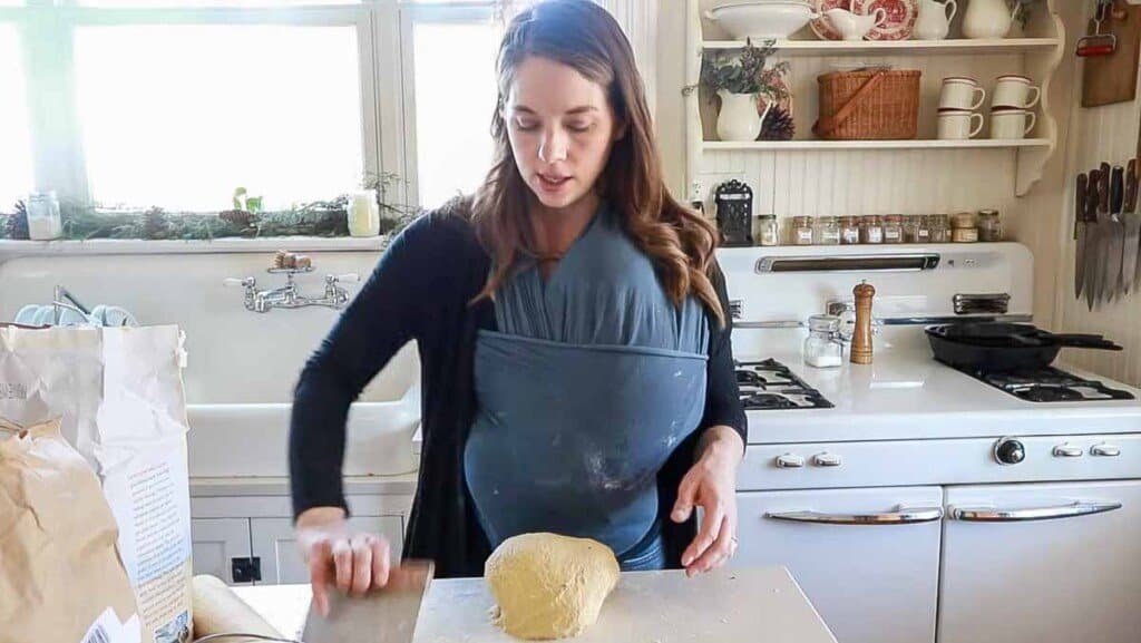 woman wearing a baby is in her white kitchen shaping a loaf of einkorn sourdough bread