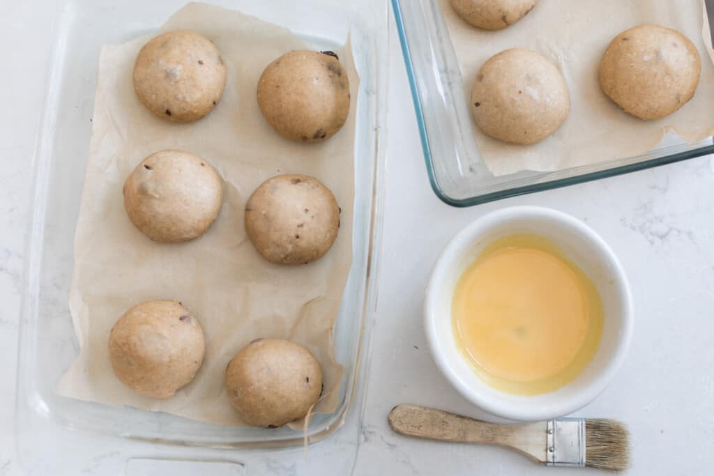 overhead photo of raw sourdough buns on two baking dishes with a white dish with a egg wash in it and a brush resting on the counter