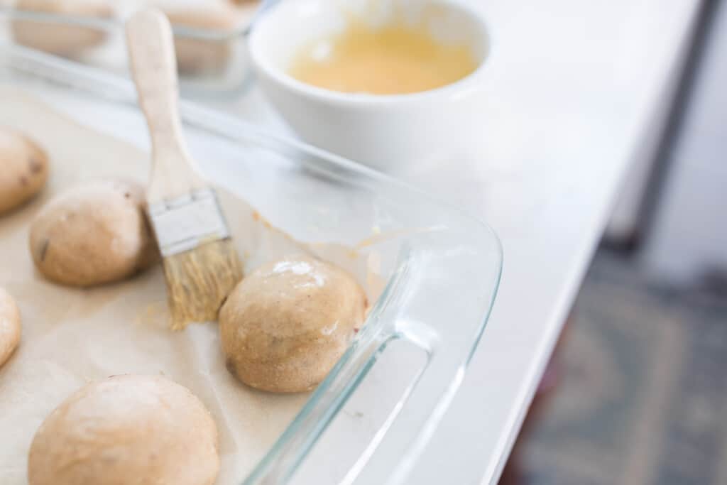 brushing on an egg wash onto sourdough hot cross buns in a glass baking dish