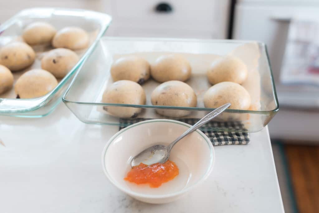 apricot jam and water in a white bowl with spoon with buns in baking dishes in behind the bowl