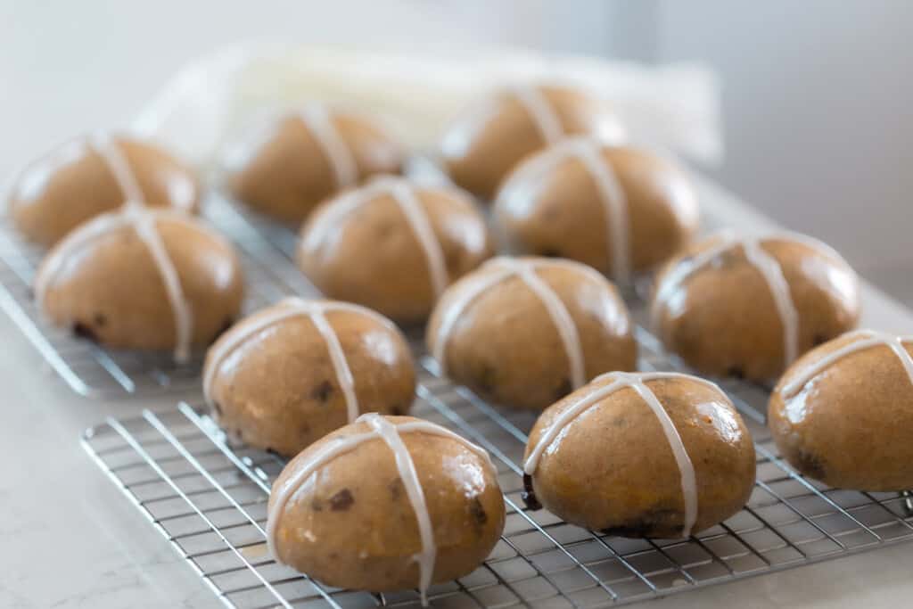 light and fluffy sourdough hot cross buns with an apricot glaze and white cross on top resting on a wire rack on a white quartz countertop