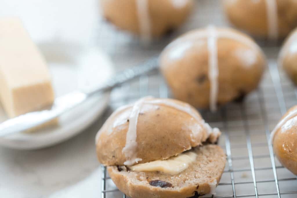 hot cross bun sliced in half with a pat of butter sandwiched between the two halves on a wire rack with other buns and a butter dish with butter in the background