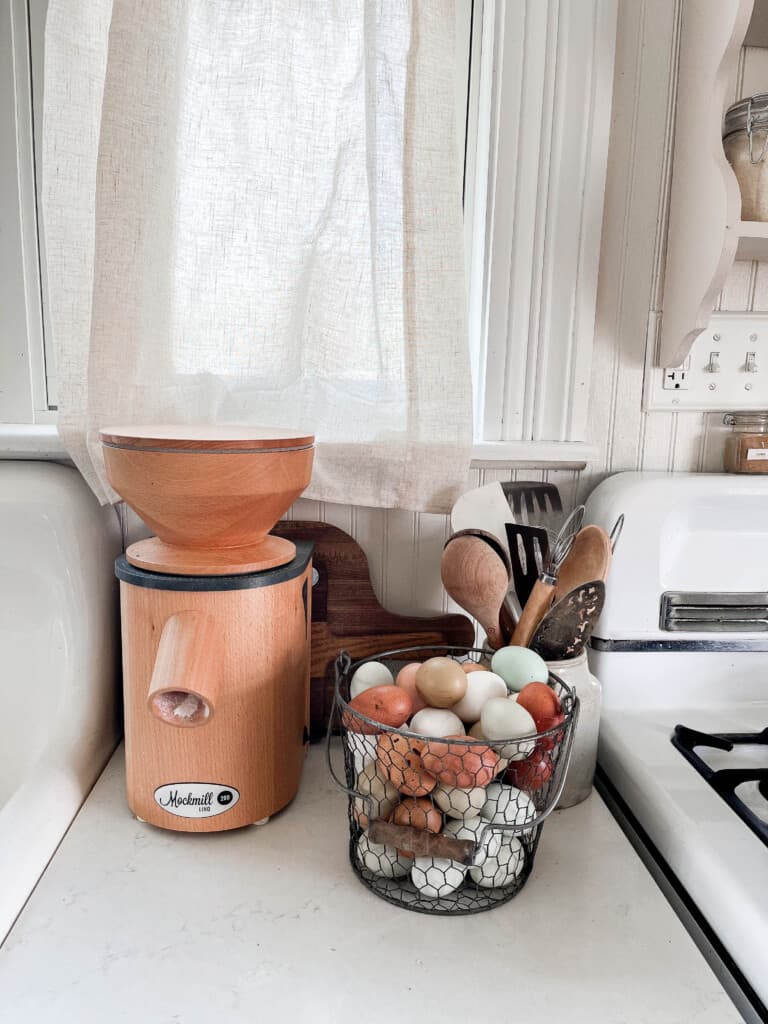 Beechwood Mockmill rests on a white countertop between a white farmhouse sink and a basket of eggs and antique stove