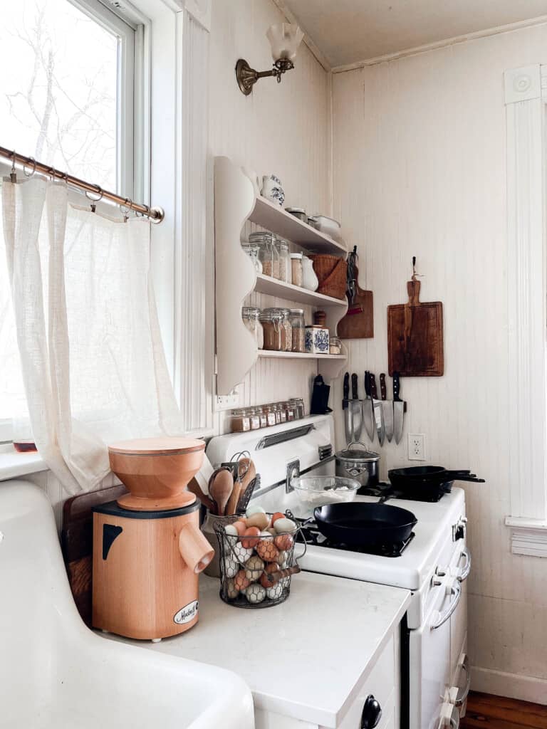 side view of a white farmhouse kitchen with a white sink, Mockmill grain mill, basket of eggs on a white countertop next to a white antique stove.