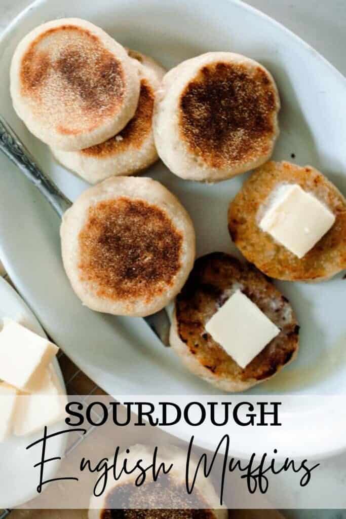 overhead photo of sourdough English muffins on a white platter. Some are sliced with pats of butter. A white dish with sliced butter sits to the left on a wire wrack over parchment paper and another English muffin in the front.