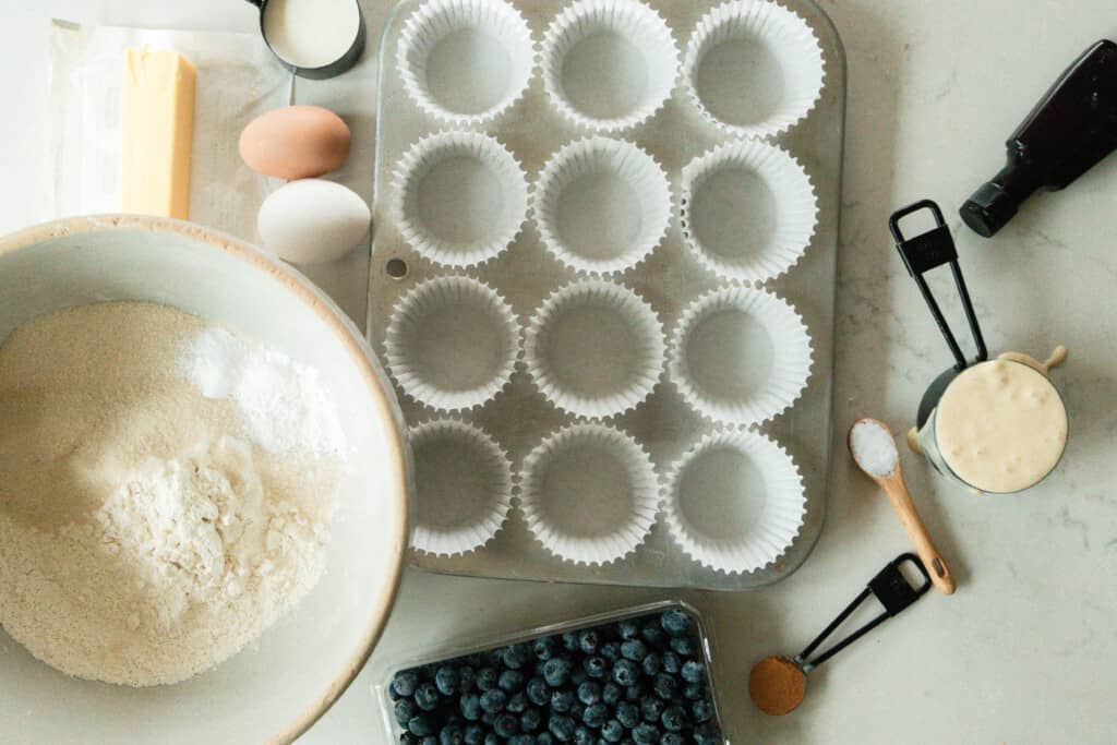 overhead photo of a muffin tray lined with muffin liners with ingredients to make sourdough blueberry muffin ingredients around the muffin tray